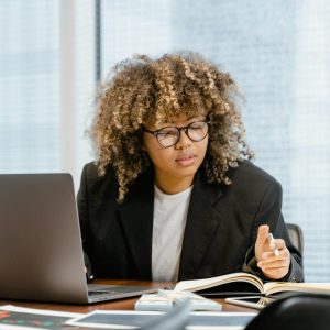 A focused woman in glasses works on a laptop and takes notes in a modern office setting.
